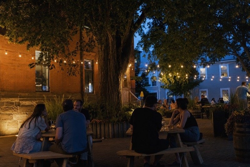 Brewery patio at dusk with picnic tables, string lights and a large elm by a red-brick wall, the kind of spot SEO Fredericton helps people find