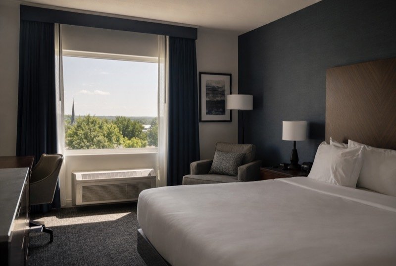 A calm hotel room with a neatly made bed, a reading chair and morning light through sheer curtains