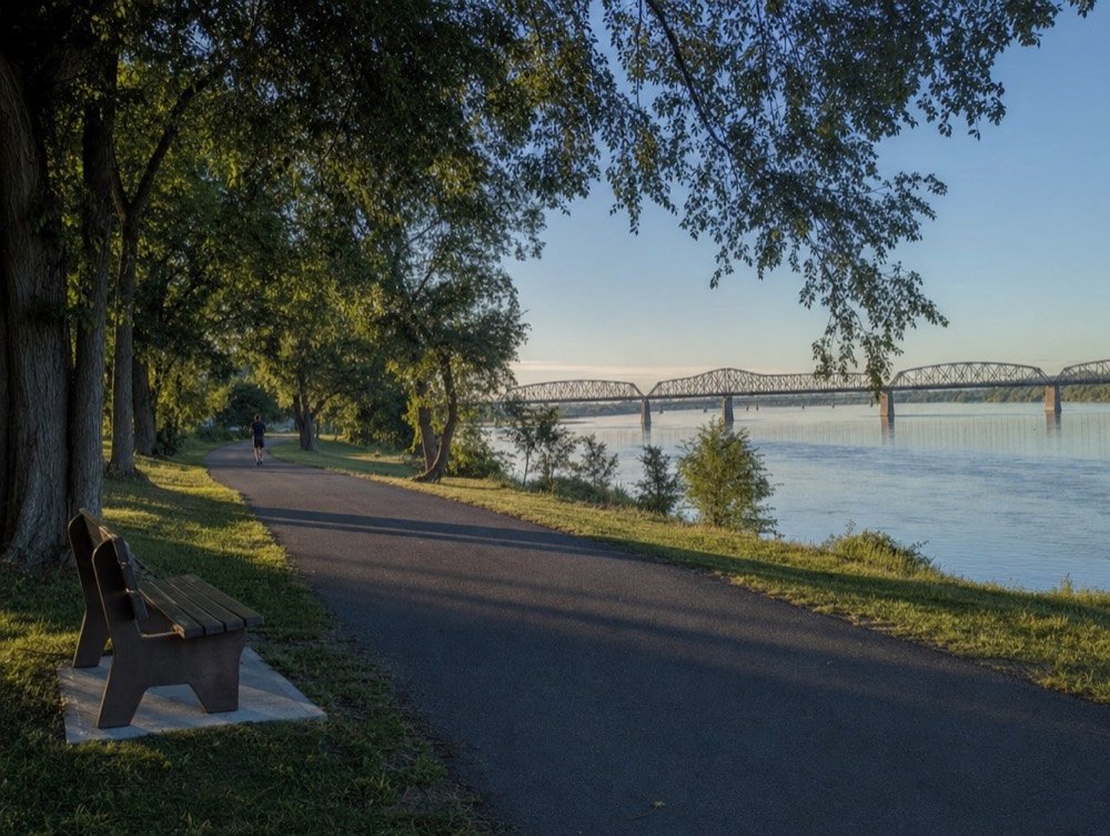 A paved riverside trail curving past a wooden bench and tall trees, with a long steel truss bridge soft in the distance and a lone jogger far down the path